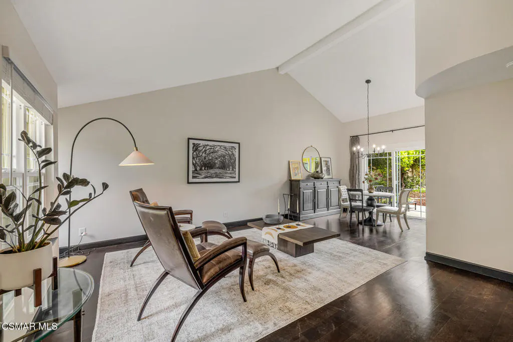 A living room with vaulted ceilings, dark wood floors, and neutral walls. Two leather chairs face a coffee table on a light rug. A dining area is visible in the background.