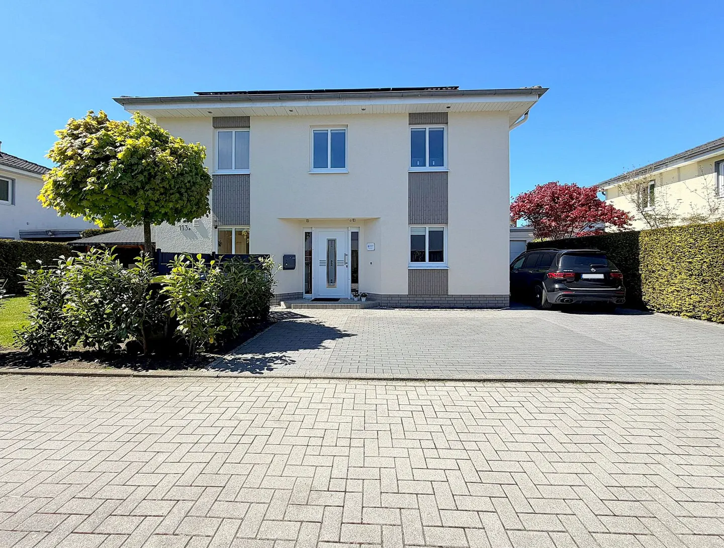 Two-story cream house with white trim, a gray brick driveway, and a black car parked in the driveway.