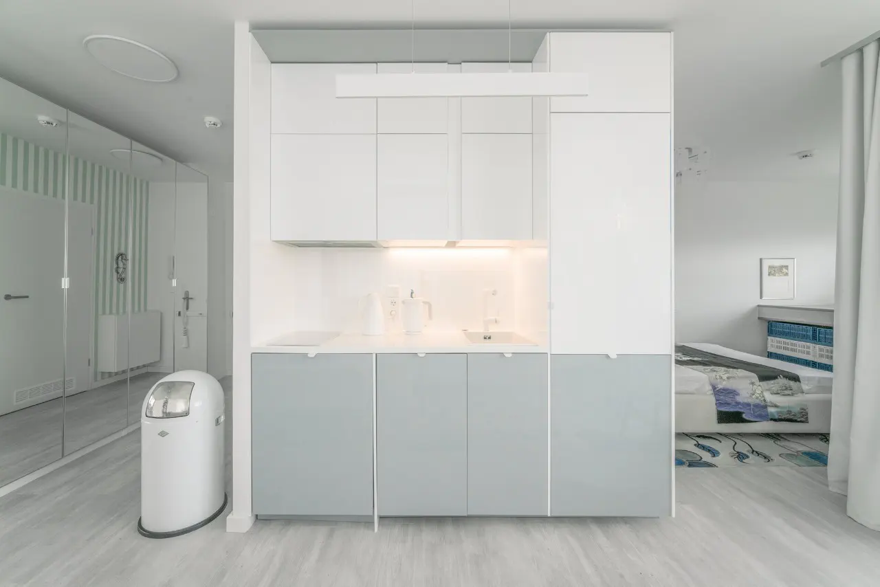 Modern kitchenette with white upper and light grey lower cabinets, white countertop, and a white trash bin in a studio room.