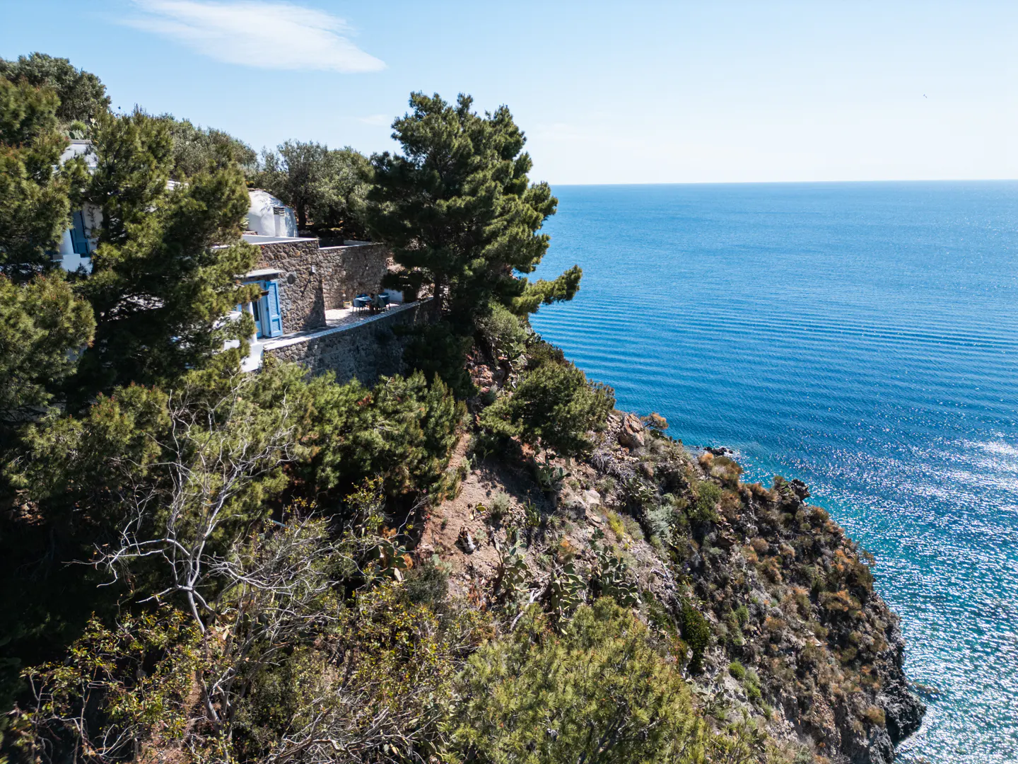 Ocean view of a white house with a blue door on a cliff surrounded by green trees under a blue sky.