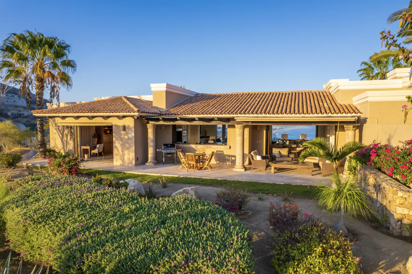 Exterior view of a tan villa with a brown tile roof, patio furniture, and lush green landscaping.