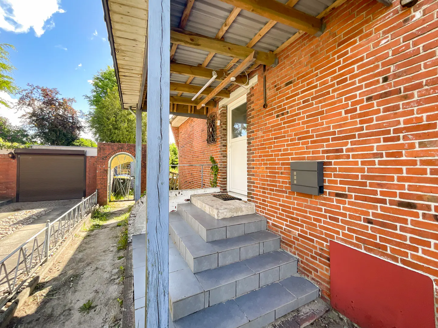 Exterior view of a red brick house with gray steps leading to a white front door under a covered porch. A gray mailbox is mounted on the brick wall.