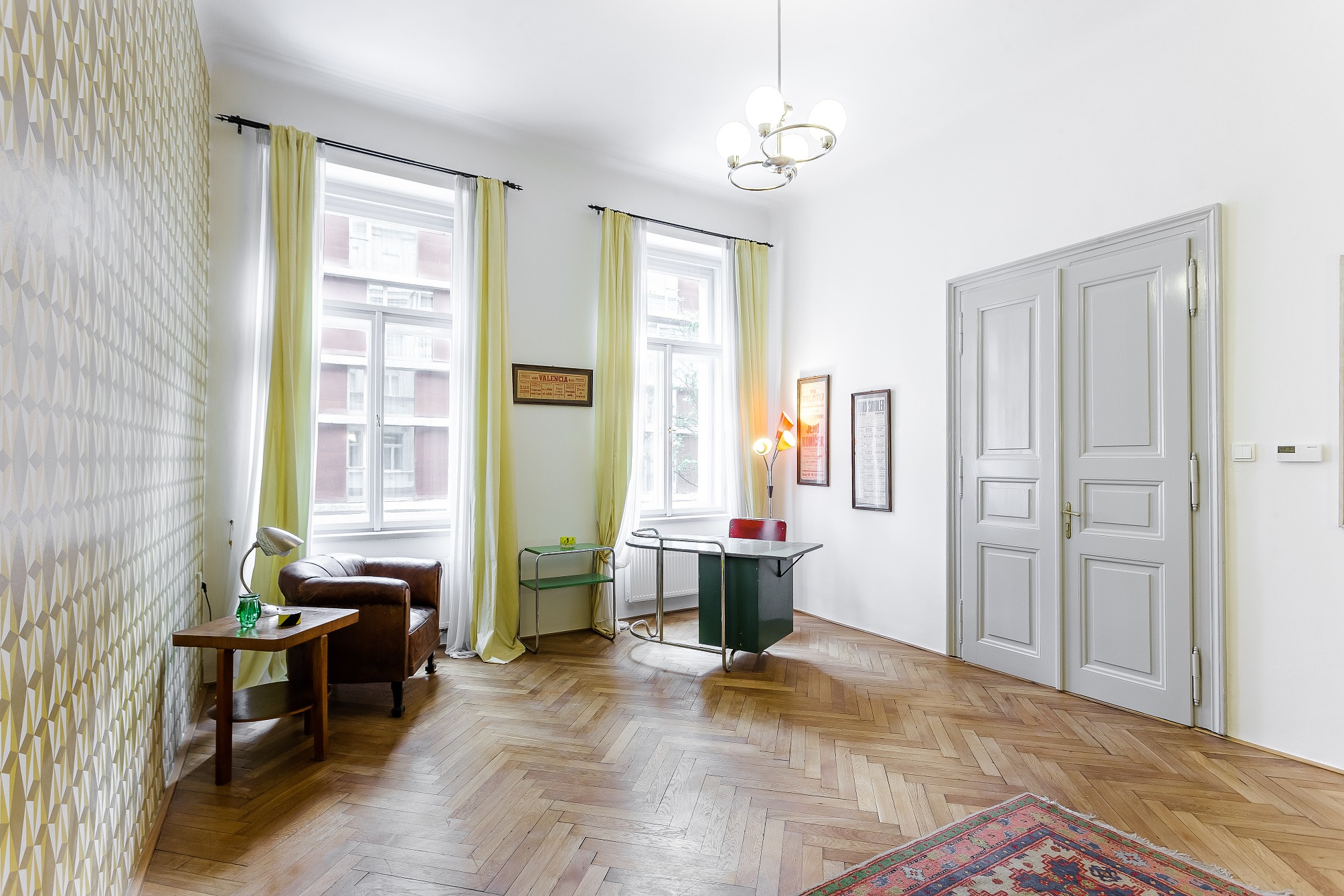 Bright room with herringbone wood floors, white walls, and patterned wallpaper. A brown leather chair sits near a window with yellow curtains. A green desk and white double doors are visible.