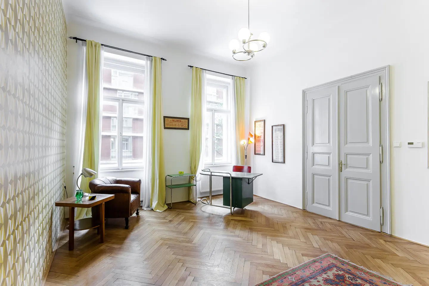 Bright room with herringbone wood floors, white walls, and patterned wallpaper. A brown leather chair sits near a window with yellow curtains. A green desk and white double doors are visible.