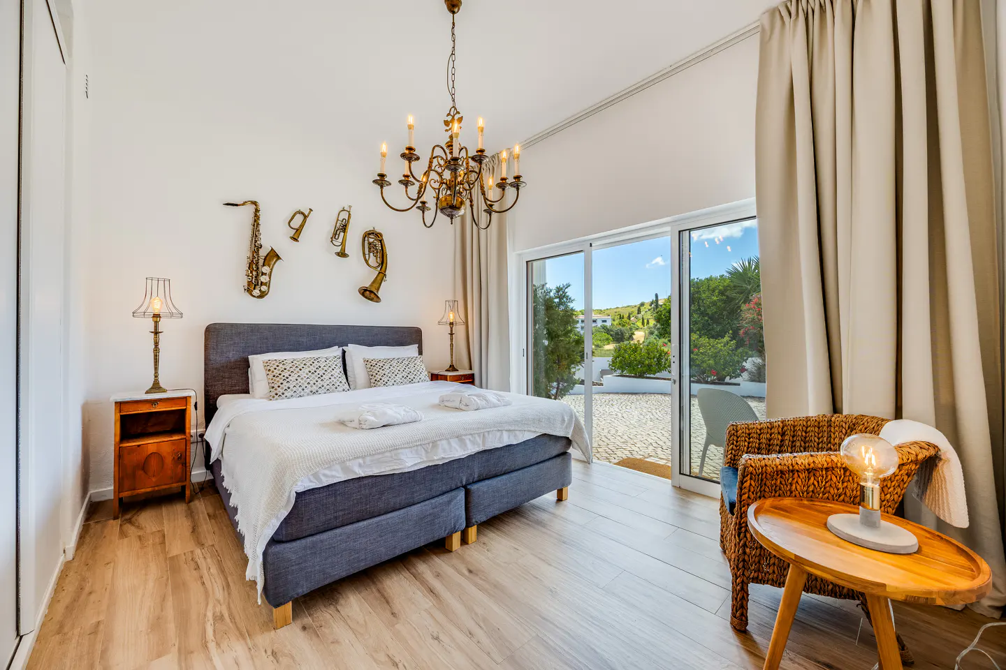 Bright bedroom with a gray bed, white linens, and musical instrument wall decor. A chandelier hangs above, and a patio is visible through glass doors.