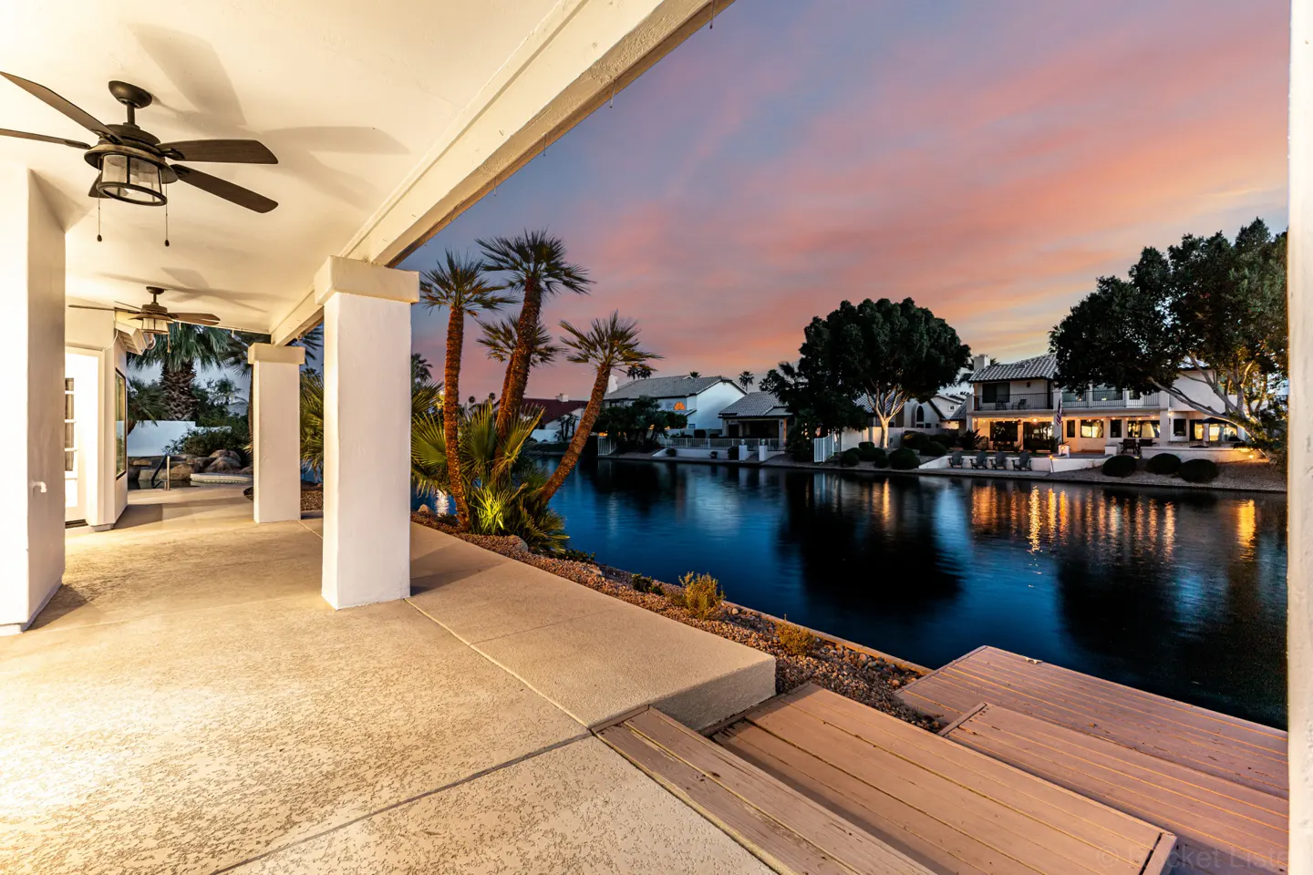Covered patio with ceiling fans overlooks a canal at sunset. Palm trees and houses line the water.