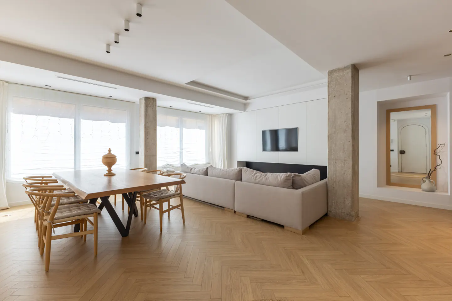 Bright, modern living and dining area with wood floors, table, and beige sofa. Large windows with white curtains provide natural light.