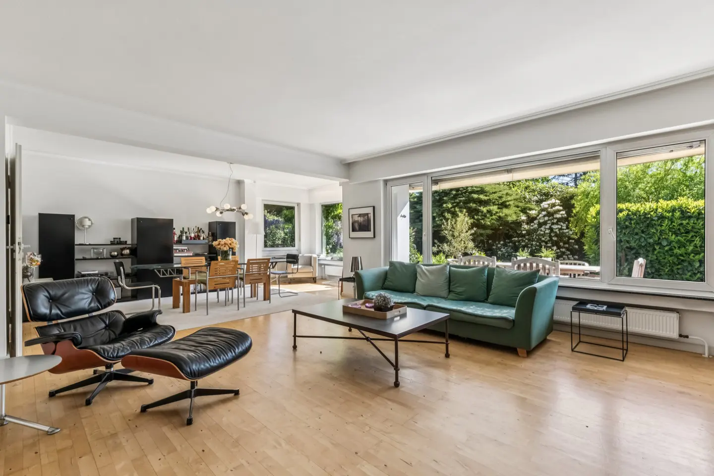 Spacious living room with a green sofa, black leather lounge chair, coffee table, and dining area overlooking a garden.