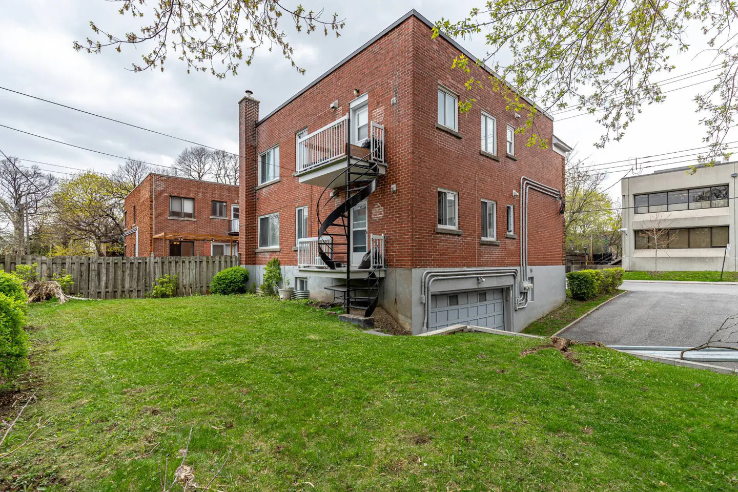 Brick apartment building with a black spiral staircase leading to a white balcony, and a green lawn in front.