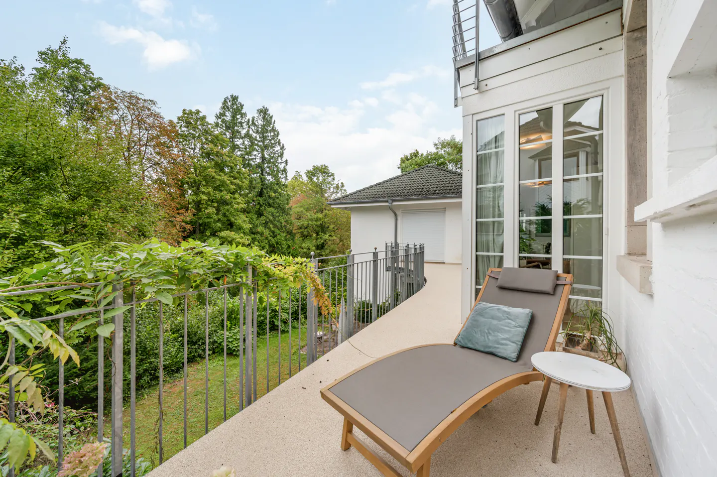 Balcony view with a gray lounge chair, blue pillow, and small white table. Green trees and a white building are visible in the background.