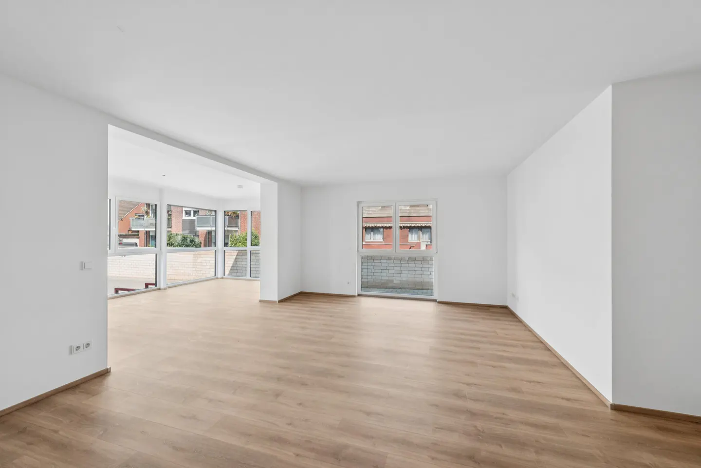 Bright, empty room with light wood floors, white walls, and large windows showing a building exterior.