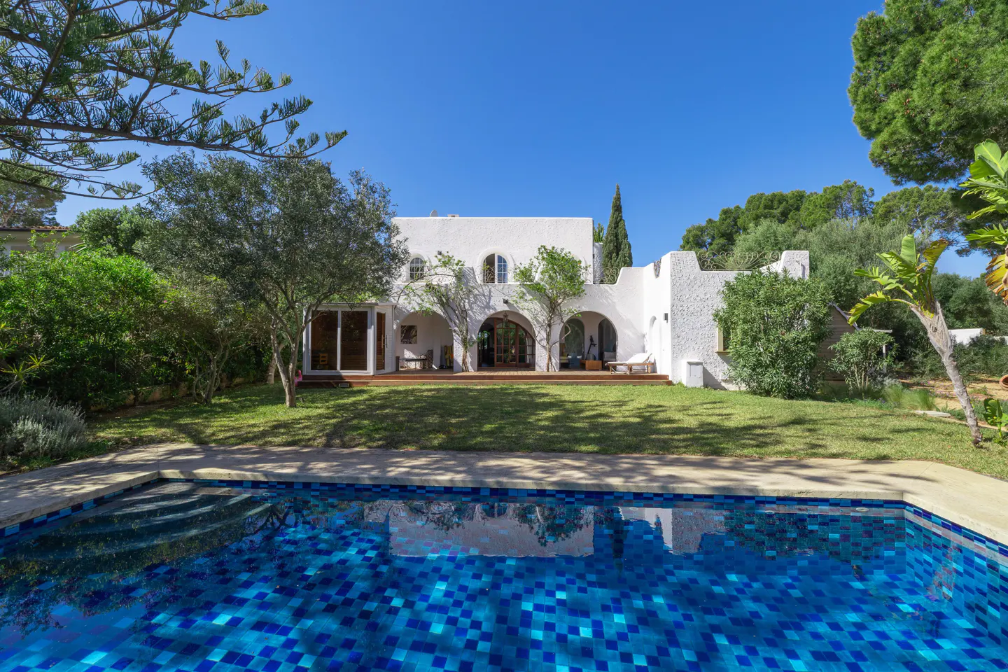 White Mediterranean-style villa with arched patio, lush green lawn, and a blue tiled swimming pool under a clear sky.