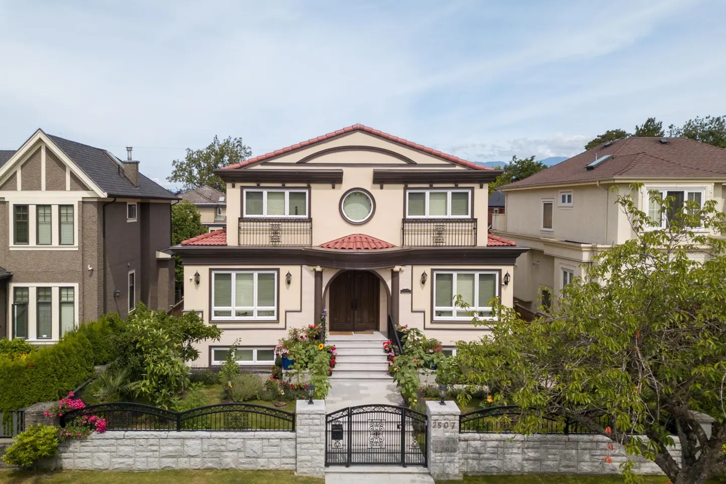Two-story beige house with a red tile roof, dark brown trim, and a stone fence with a black metal gate.