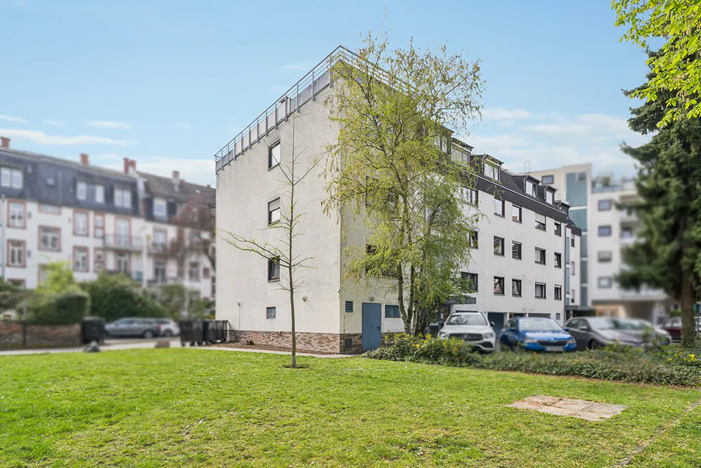 Exterior view of a white apartment building with a black roof, surrounded by green grass and trees, with cars parked nearby.