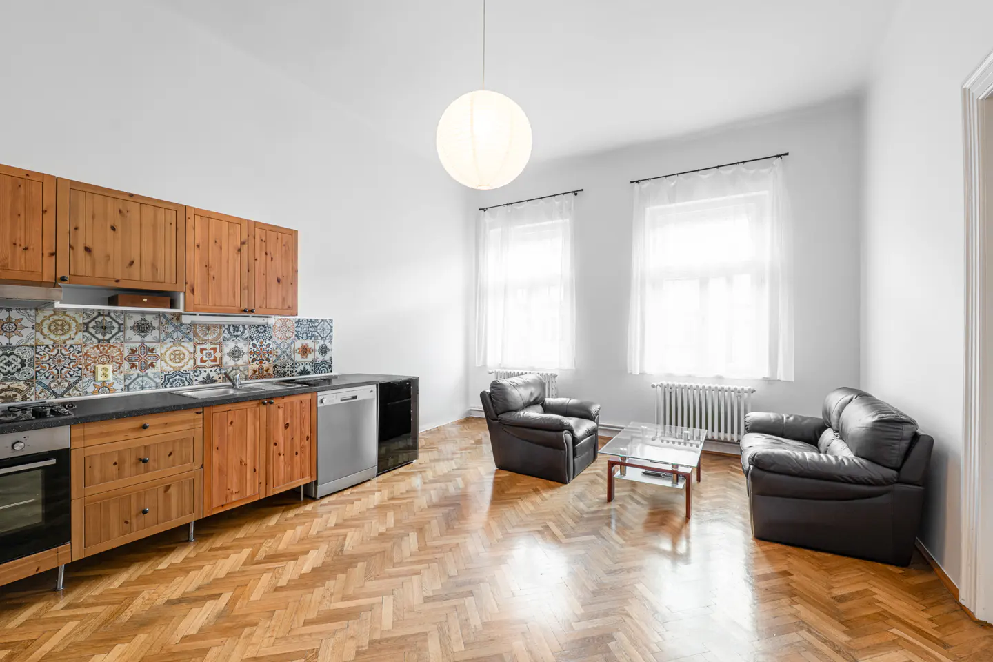 Bright open-plan kitchen and living area with wooden cabinets, patterned tile backsplash, and two black leather sofas.