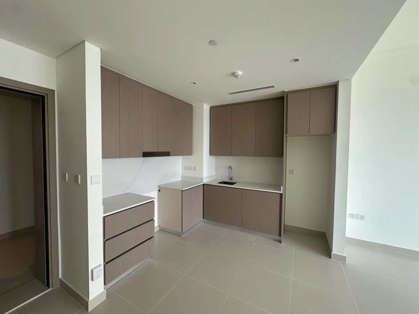 A modern, empty kitchen with brown cabinets, white countertops, and gray tile flooring. A doorway is visible on the left.