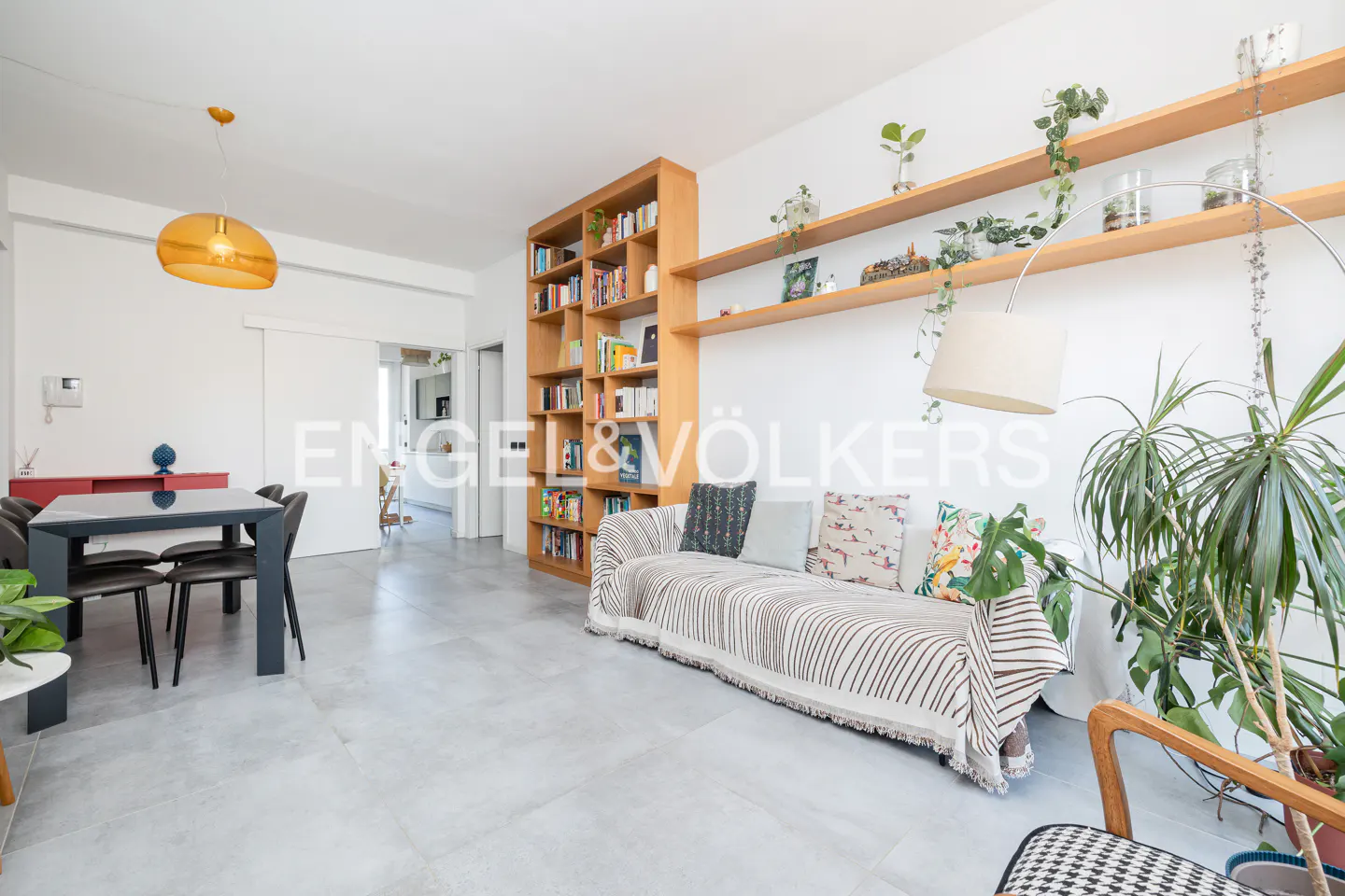 Bright living room with a sofa, wooden bookshelf, and shelves with plants. A dining table is visible in the background.