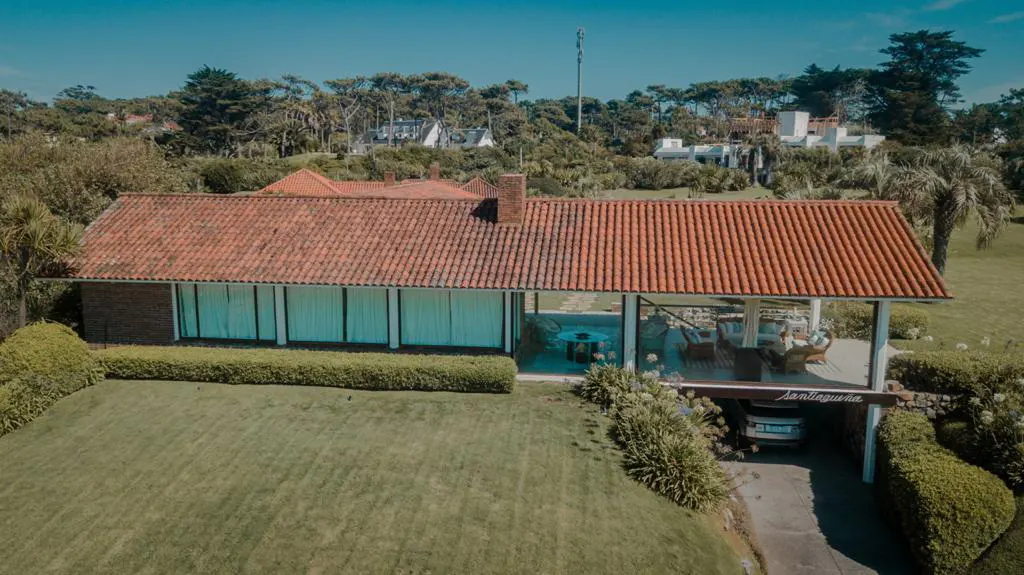 Aerial view of a single-story house with a red tile roof, large windows, and a covered patio with a car parked underneath. Green lawn and trees surround the house.