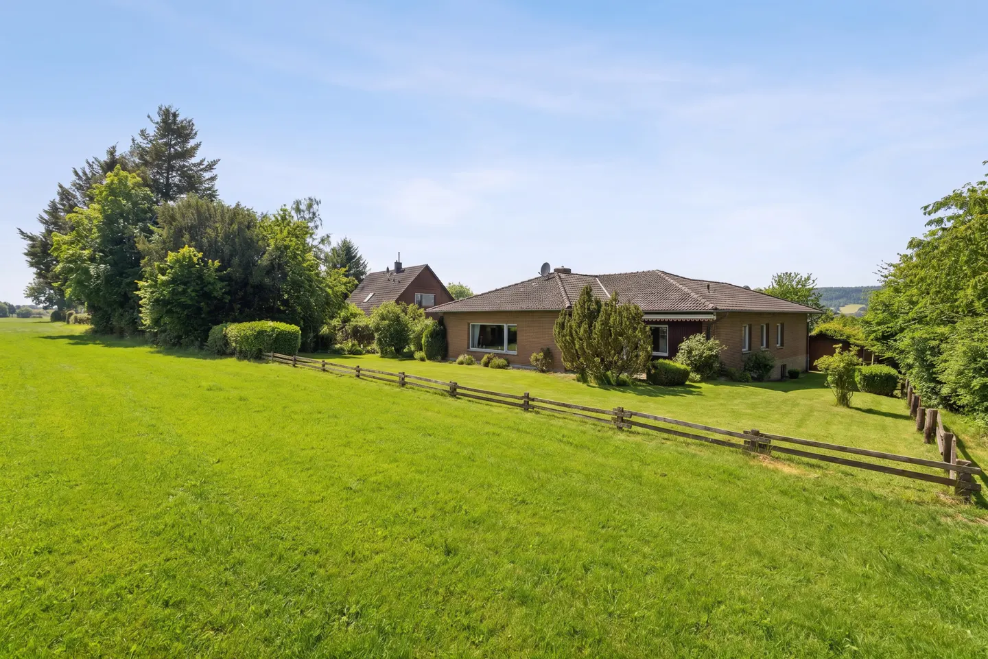A single-story tan house with a gray roof sits on a green lawn behind a wooden fence under a blue sky.