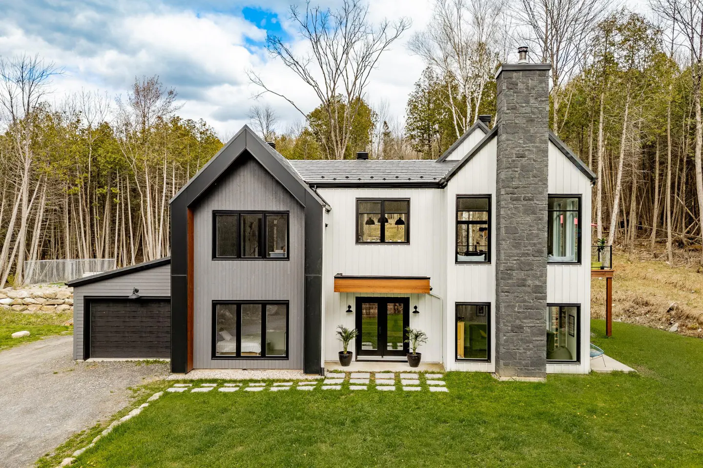 Two-story modern farmhouse with gray and white siding, black trim, and a stone chimney, set on a green lawn with trees in the background.