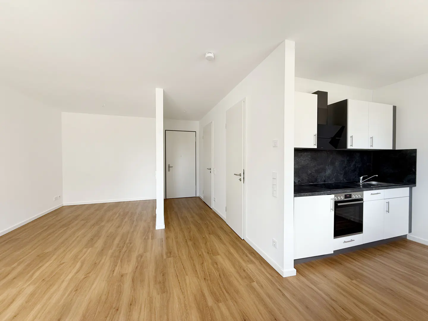 Bright, empty apartment with wood floors, white walls, and a modern kitchen with black countertops.