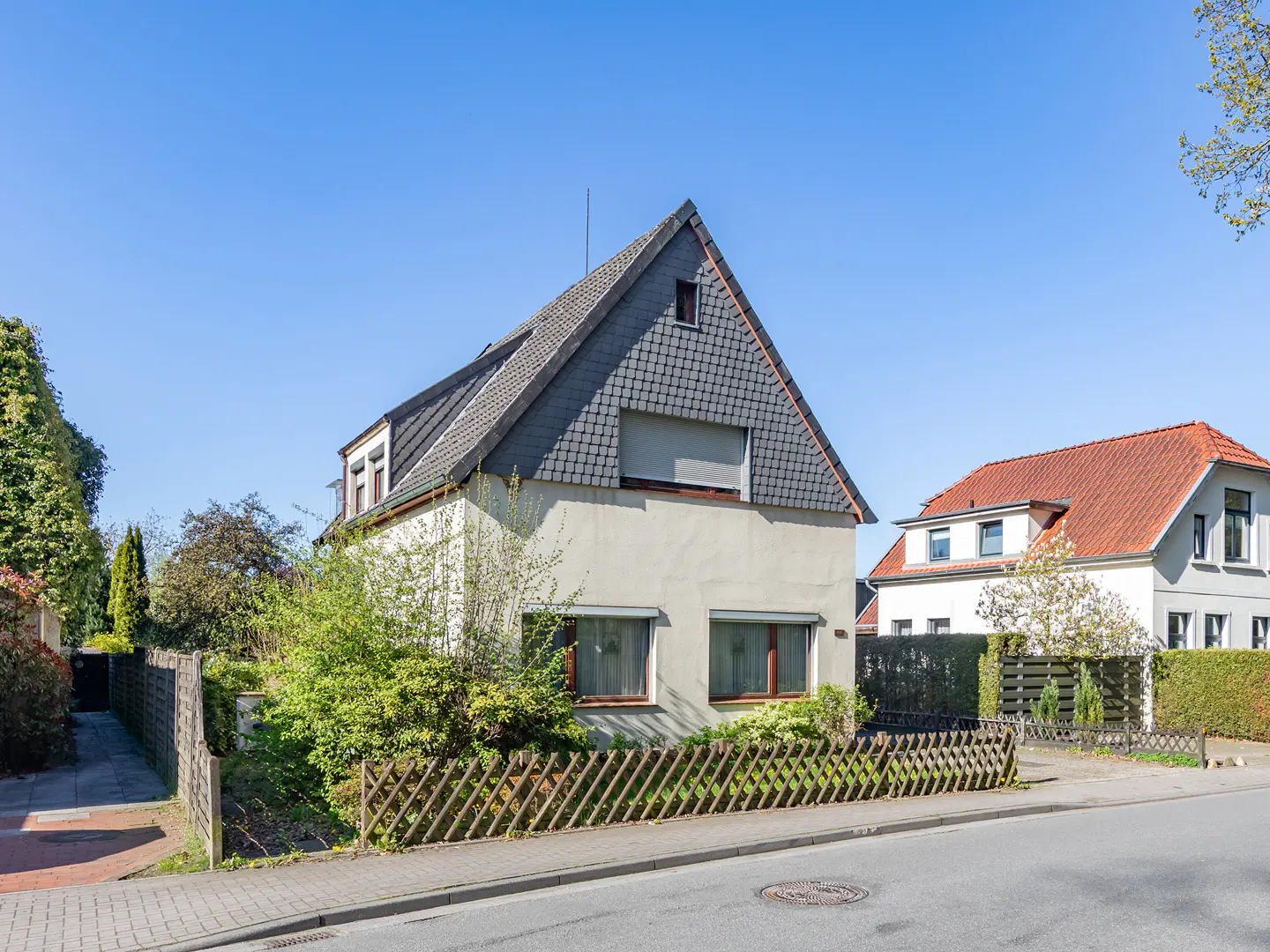 Two-story house with a gray roof and light-colored walls, surrounded by a wooden fence and greenery. Clear blue sky in the background.