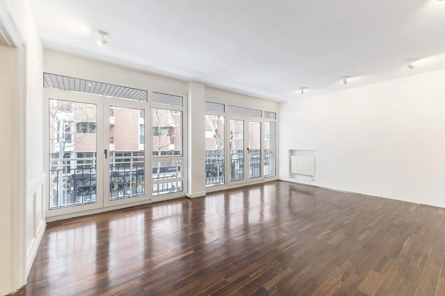 Bright, empty room with dark wood floors and white walls. Large windows with white frames overlook a city street.