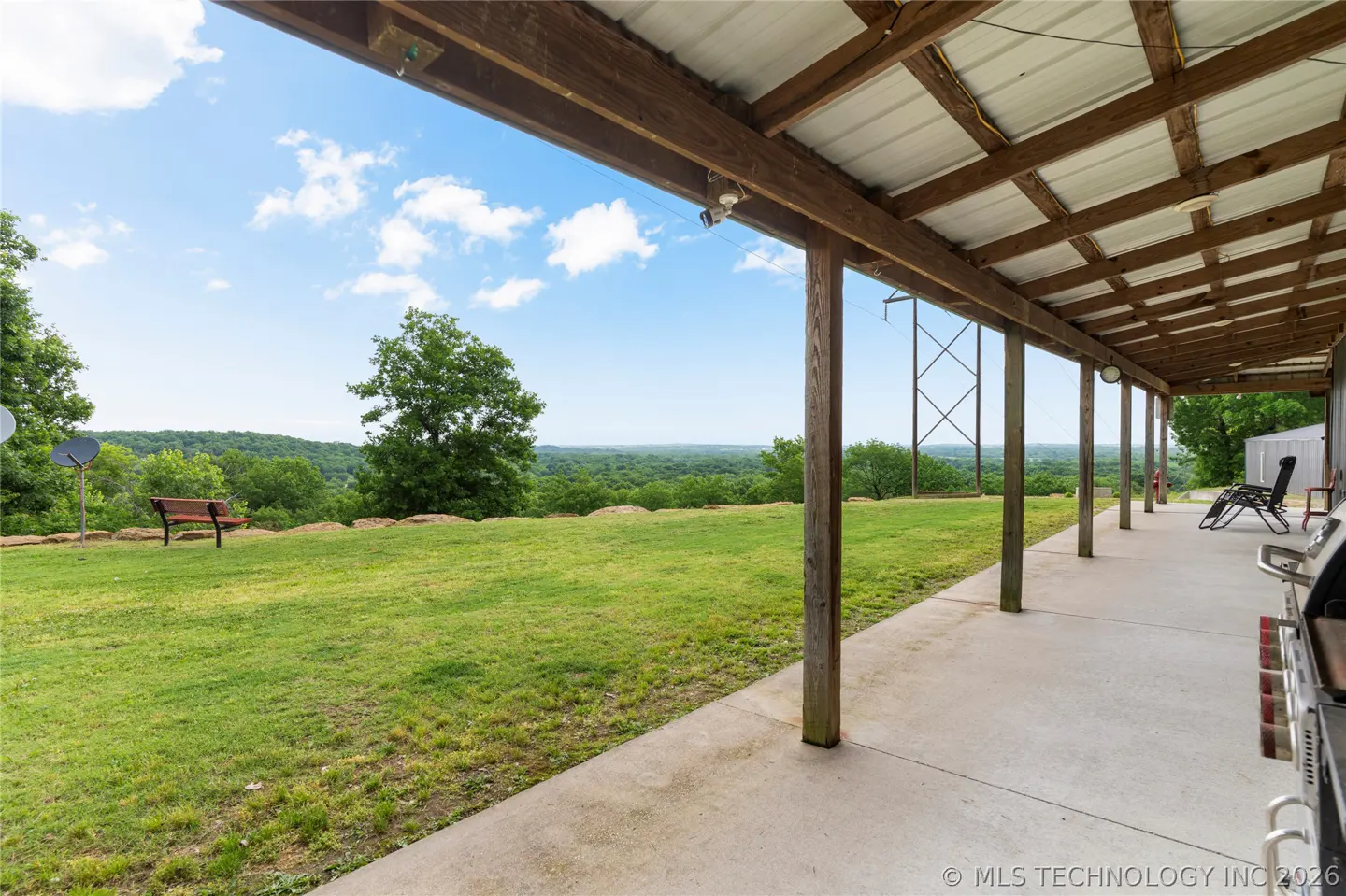 Covered patio with wooden beams overlooks a green lawn and distant trees under a blue sky. A bench sits on the lawn.