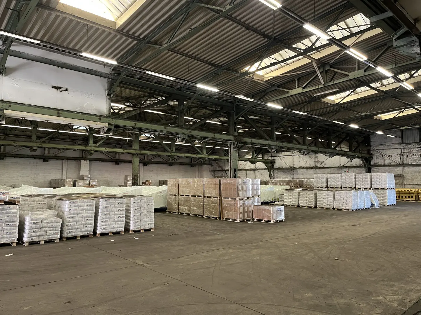 A large warehouse interior with stacks of wrapped pallets under a high, metal-beamed ceiling.