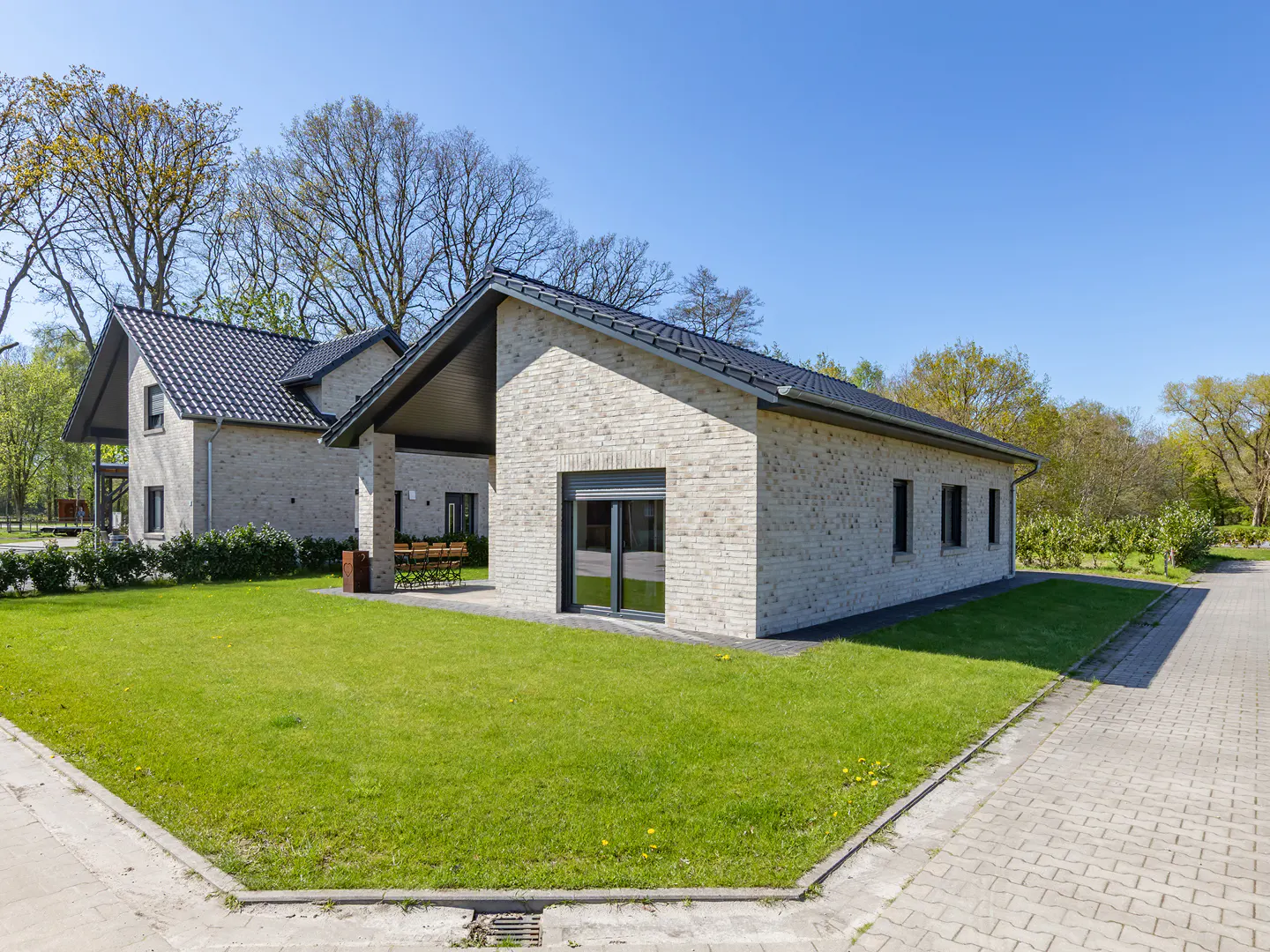 Two modern, light brick homes with dark roofs and trim, surrounded by green lawns and trees under a clear blue sky.