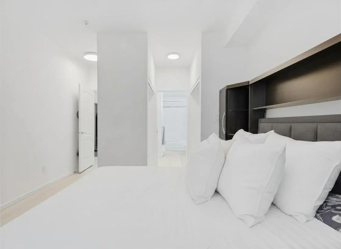 A bright bedroom with white walls, a white bed with white pillows, and a dark headboard with shelves. A hallway leads to a closet.