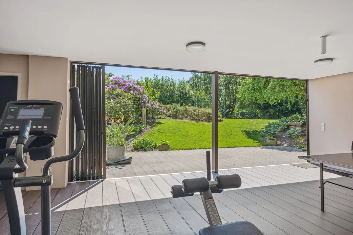 Indoor gym with exercise equipment and a ping pong table. Sliding glass doors open to a green lawn and garden.