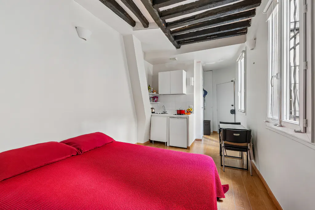 A studio apartment with a red bed, white walls, and a kitchenette. Exposed dark wood beams are on the ceiling. Three windows let in natural light.