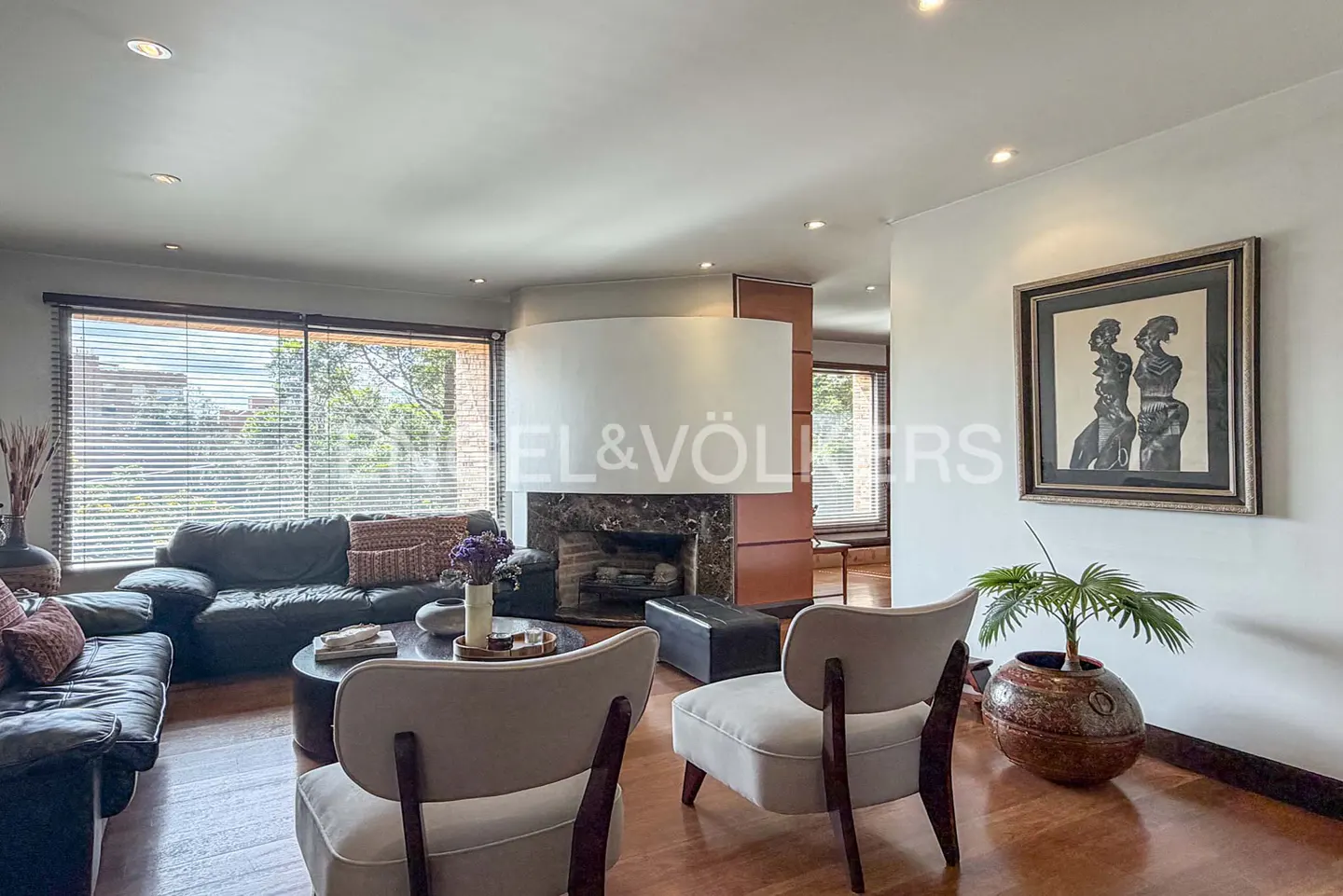 Modern living room with dark leather sofas, two light-colored armchairs, a central fireplace, and framed wall art.