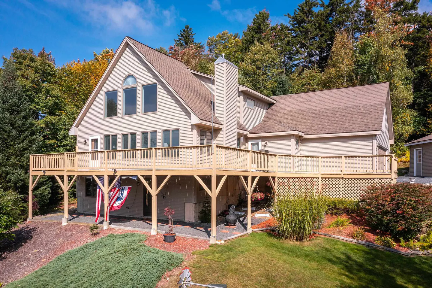 Beige two-story house with a large wooden deck, chimney, and an American flag hanging from the deck. Trees in the background.