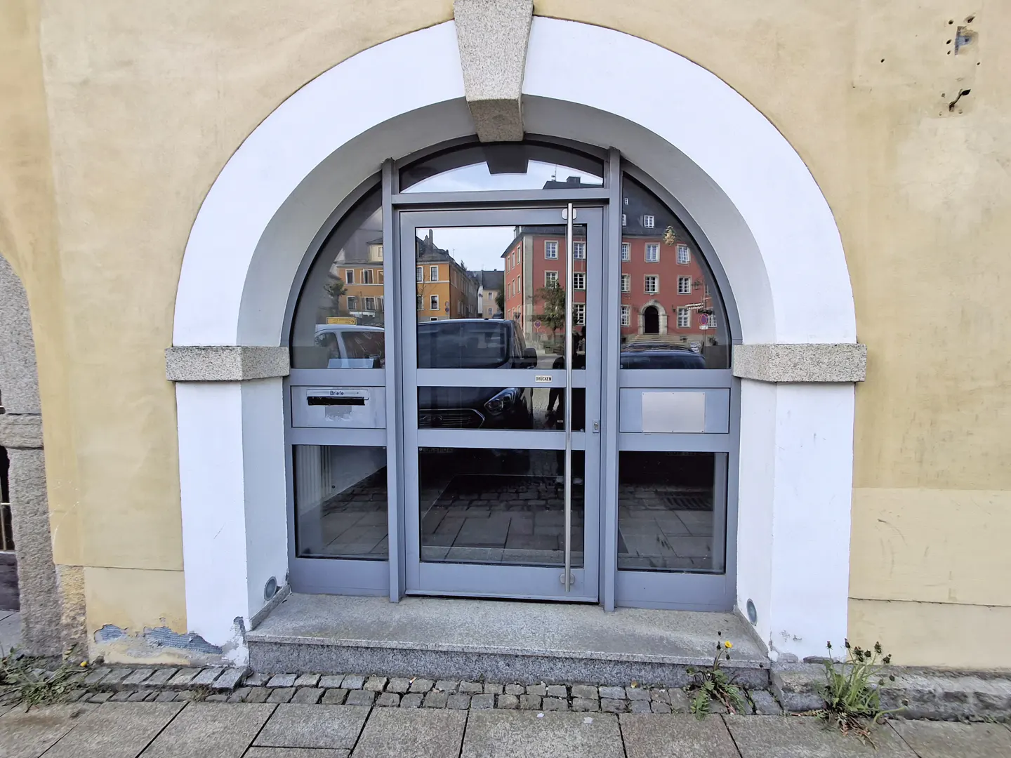 Arched doorway with gray door and white trim on a tan building. The door reflects buildings and cars on the street.