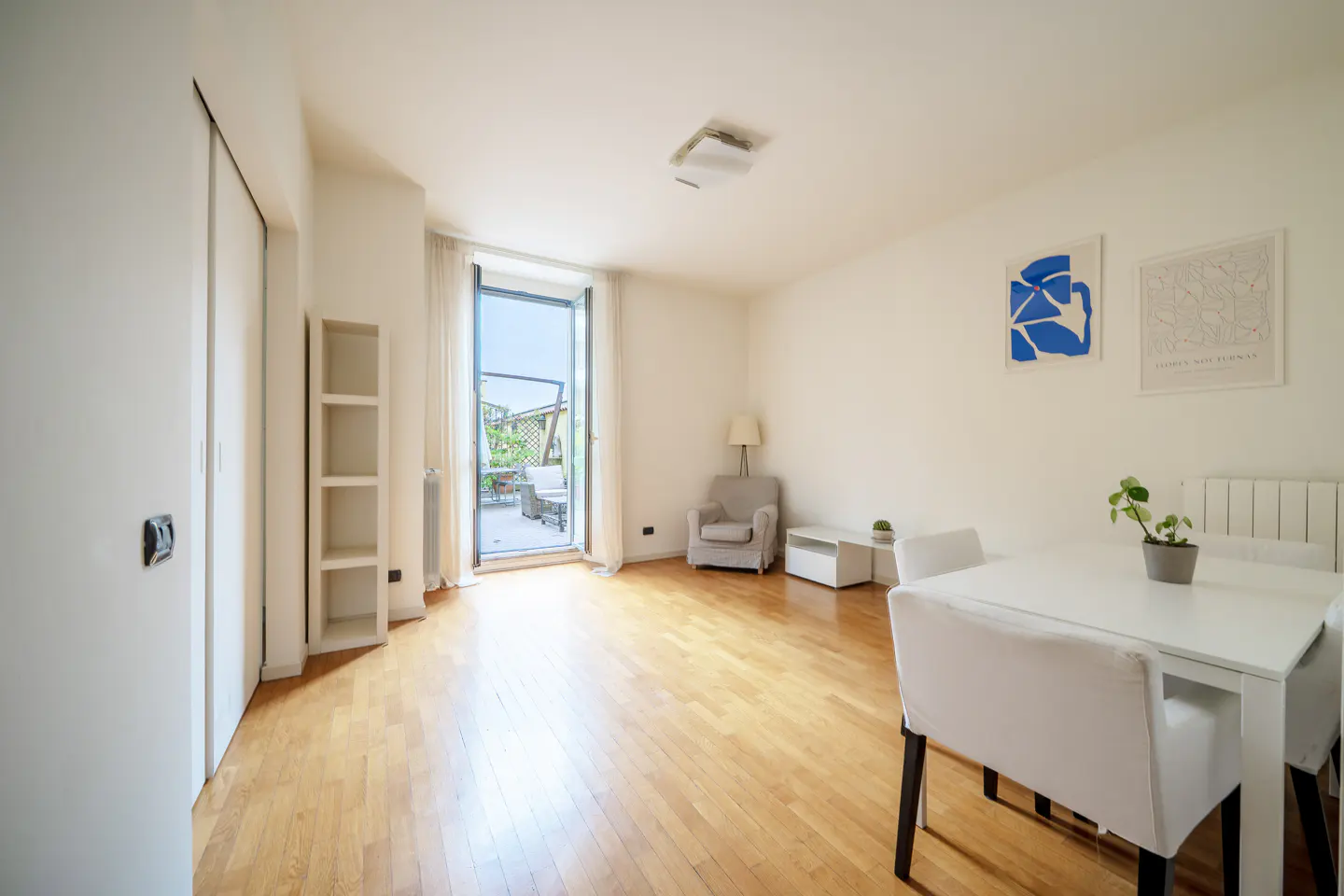 Bright living room with wood floors, white walls, and a balcony. A white table and chairs are in the foreground. Artwork hangs on the wall.