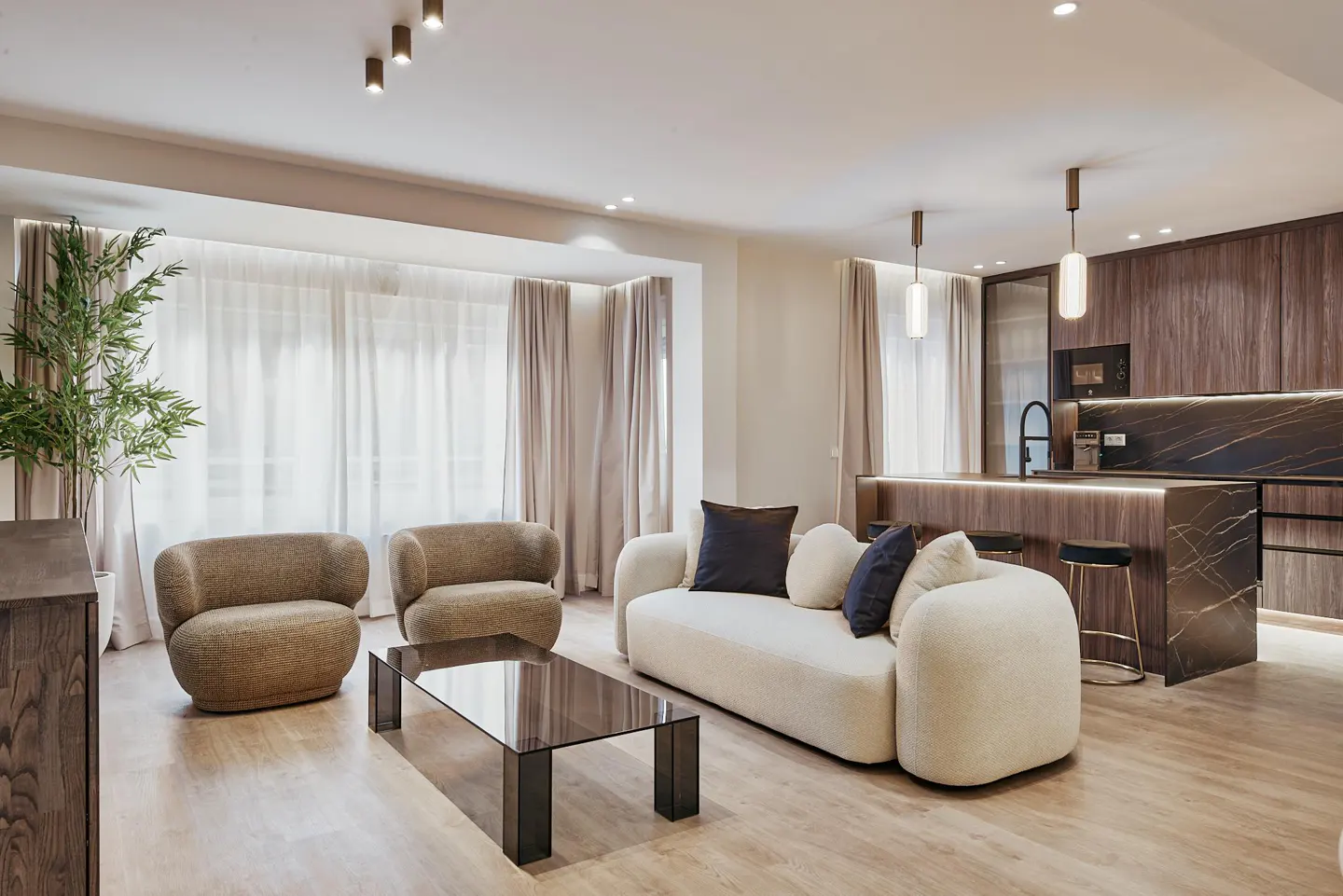 A modern living room with two brown chairs, a white sofa with pillows, and a glass coffee table. The kitchen is in the background.