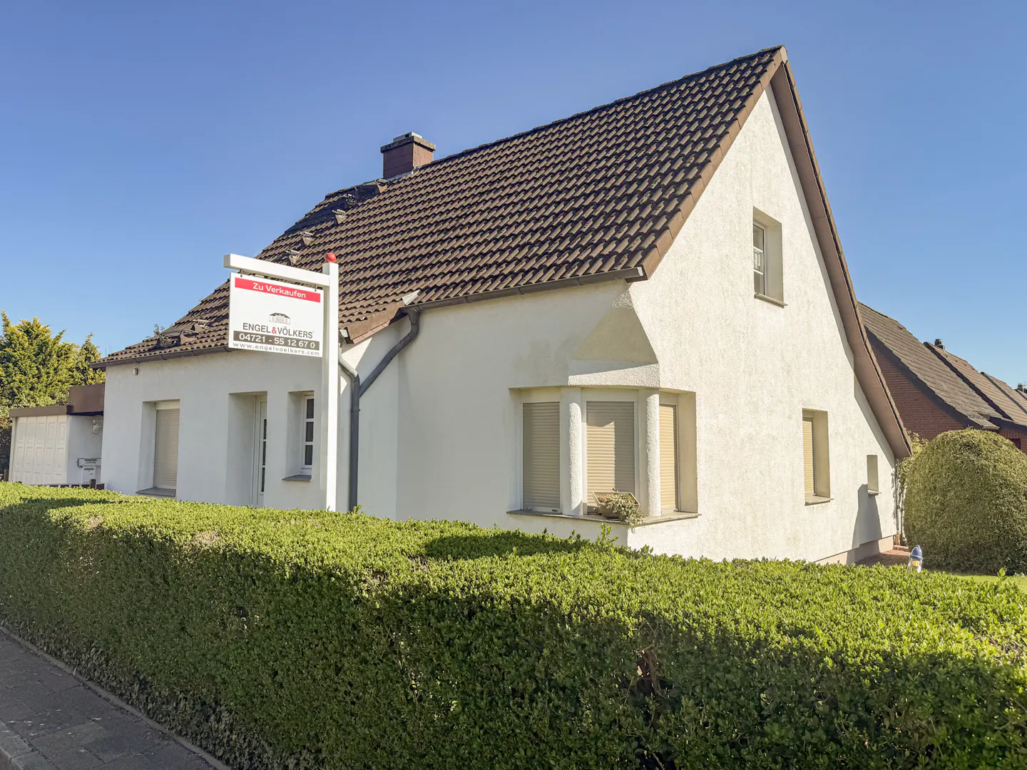 A white house with a brown roof and an Engel & Völkers "For Sale" sign in front.