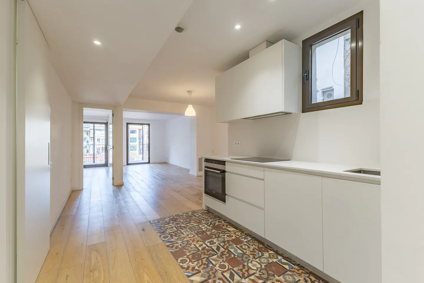 Bright, modern apartment interior with white walls, wood floors, and a white kitchen. A patterned tile rug adds color. Balconies are visible in the background.