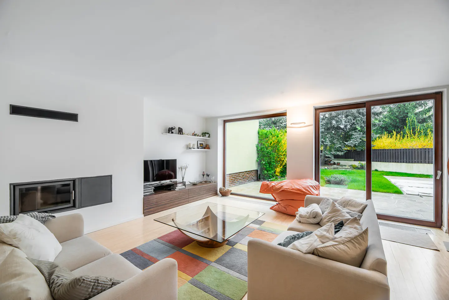 Bright living room with white walls, beige sofas, and a glass coffee table on a colorful rug. Large windows overlook a green backyard.