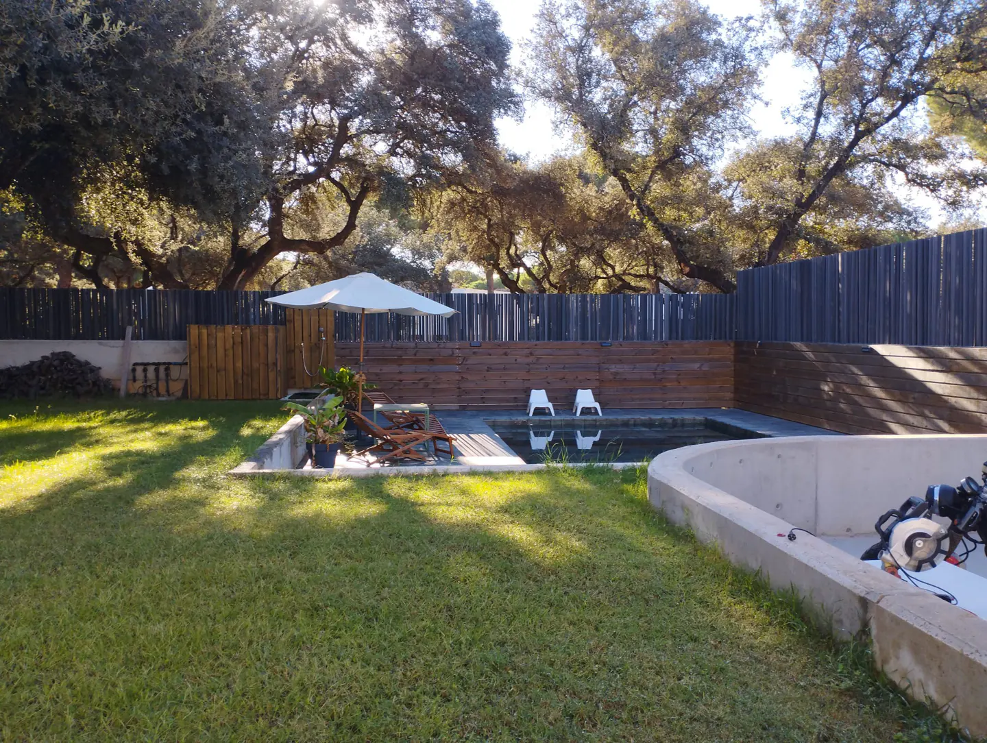 Backyard view with a pool, lounge chairs, and a white umbrella. Trees and a wooden fence surround the green lawn.
