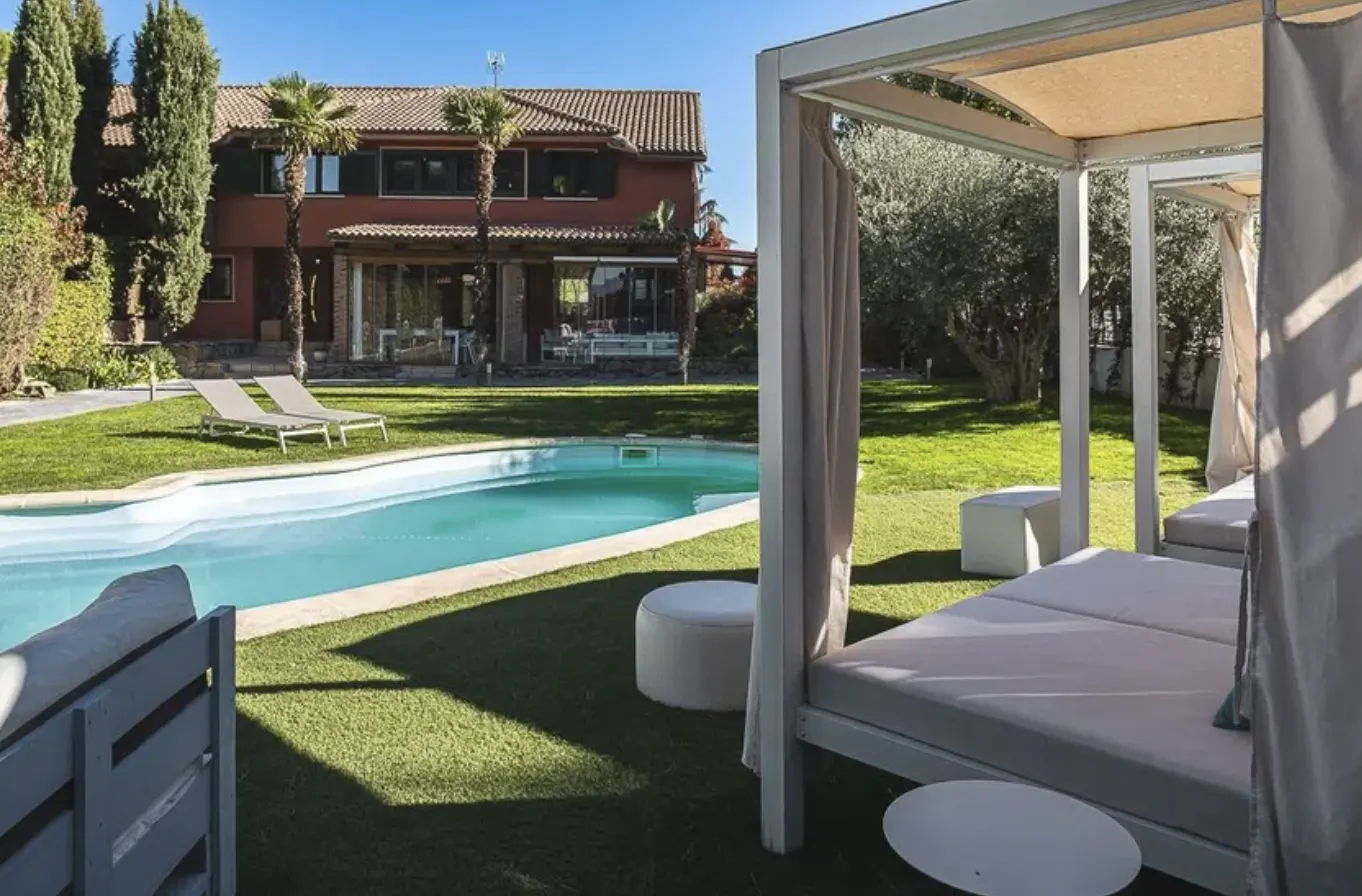 A backyard with a pool, lounge chairs, and a white canopy bed. A red house is in the background.