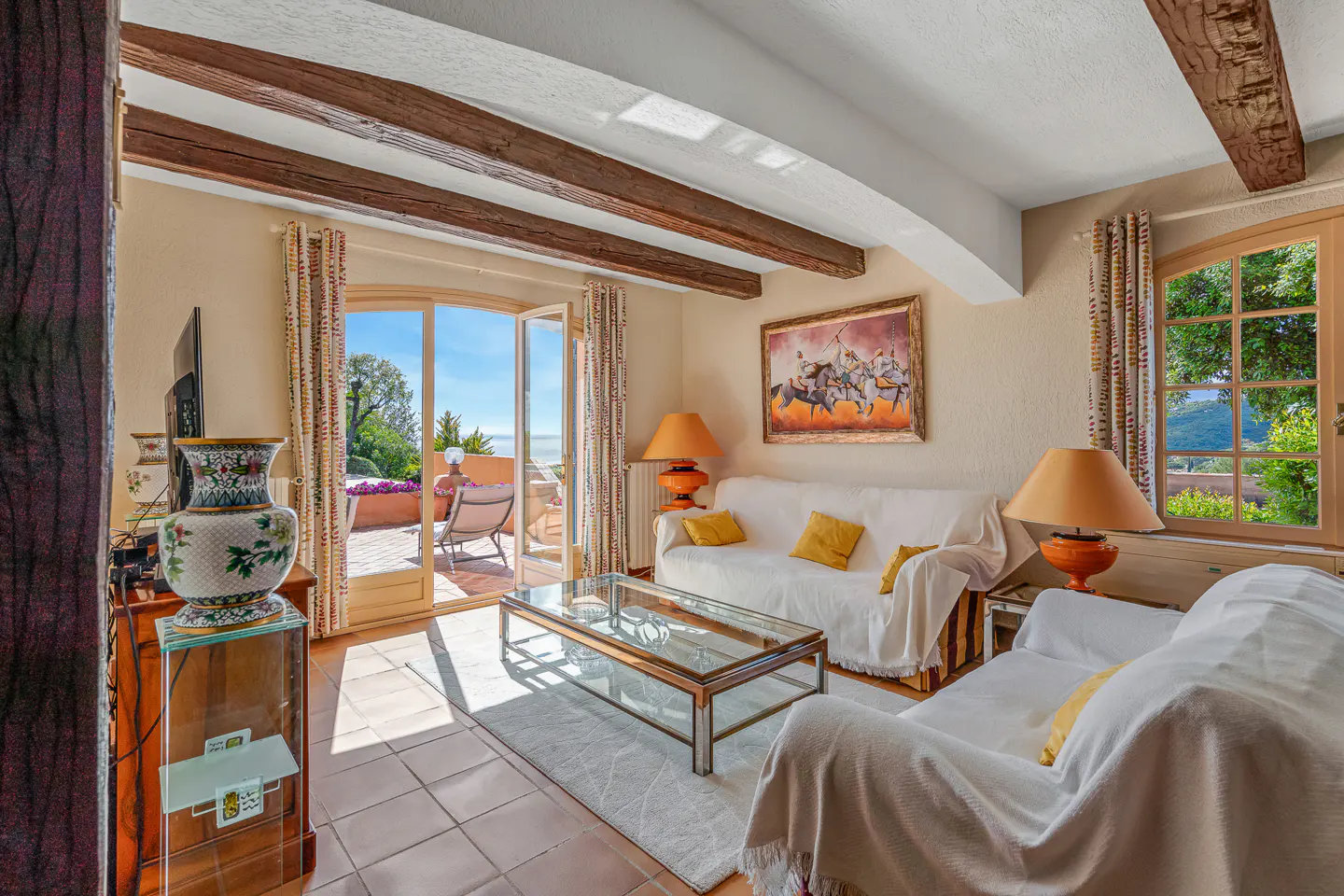 A bright living room with white sofas, yellow pillows, and a glass coffee table. French doors open to a sunny patio with ocean views.