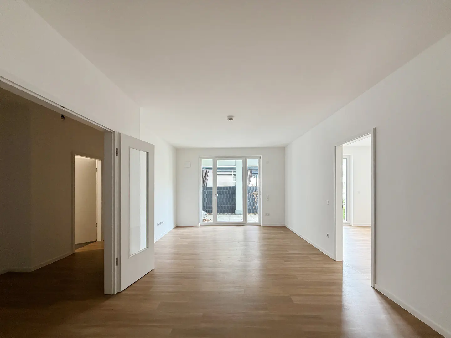 Bright, empty room with light wood floors and white walls. Glass doors lead to a balcony. Two doorways are visible.