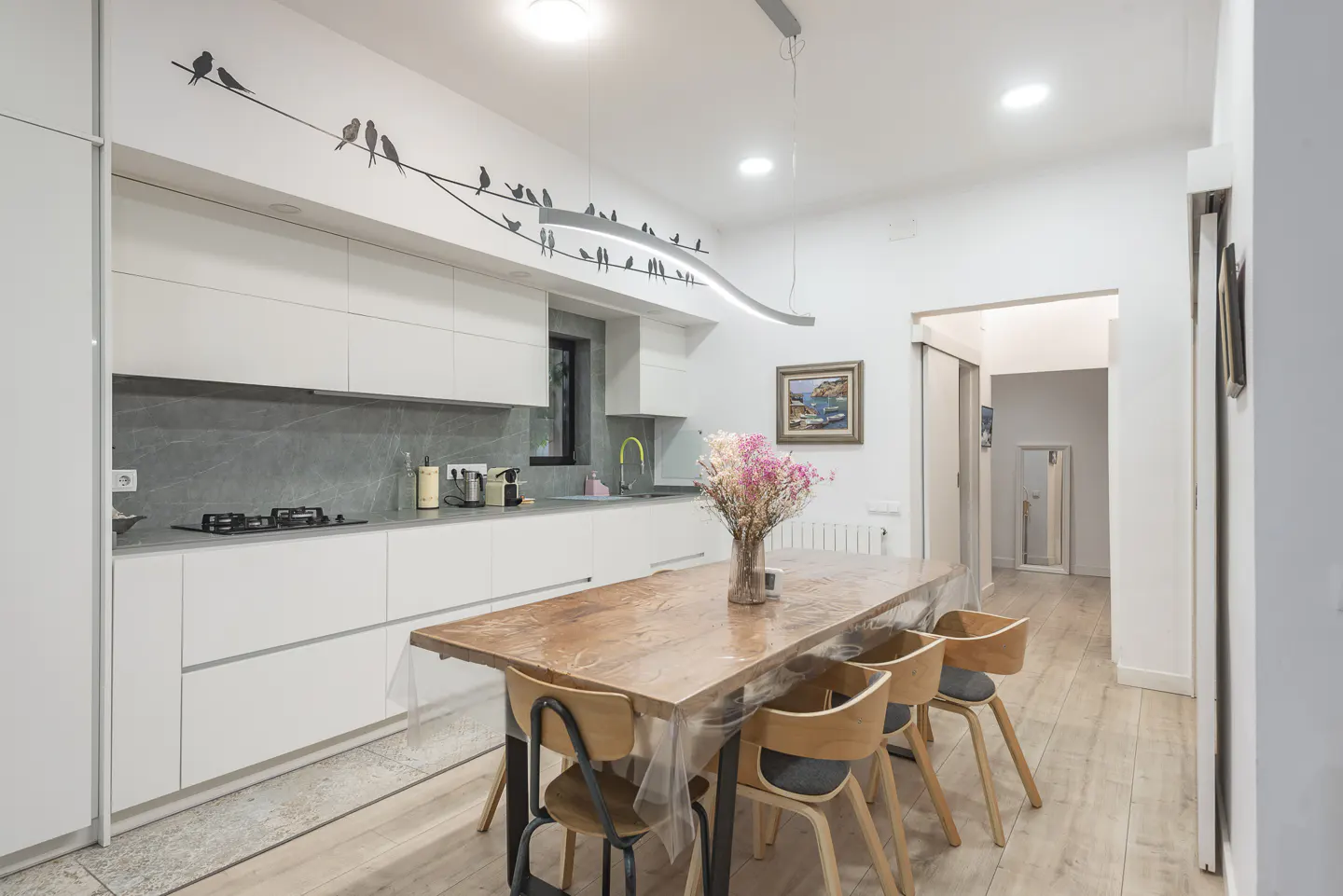 Modern white kitchen with a wooden dining table, four chairs, and a wall decal of birds on a wire.