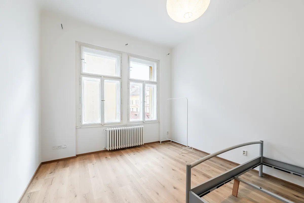 Bright, empty room with white walls, wood floor, and a large window. A radiator sits beneath the window, and a metal bed frame is in the foreground.