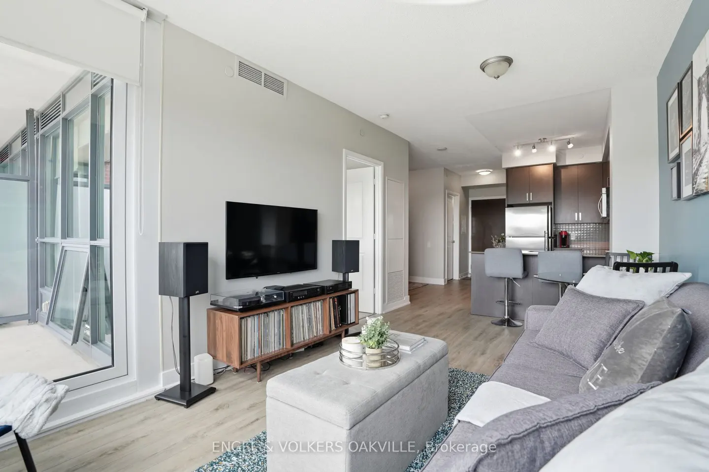 A modern condo living room with a gray sofa, TV, speakers, and an open kitchen in the background.