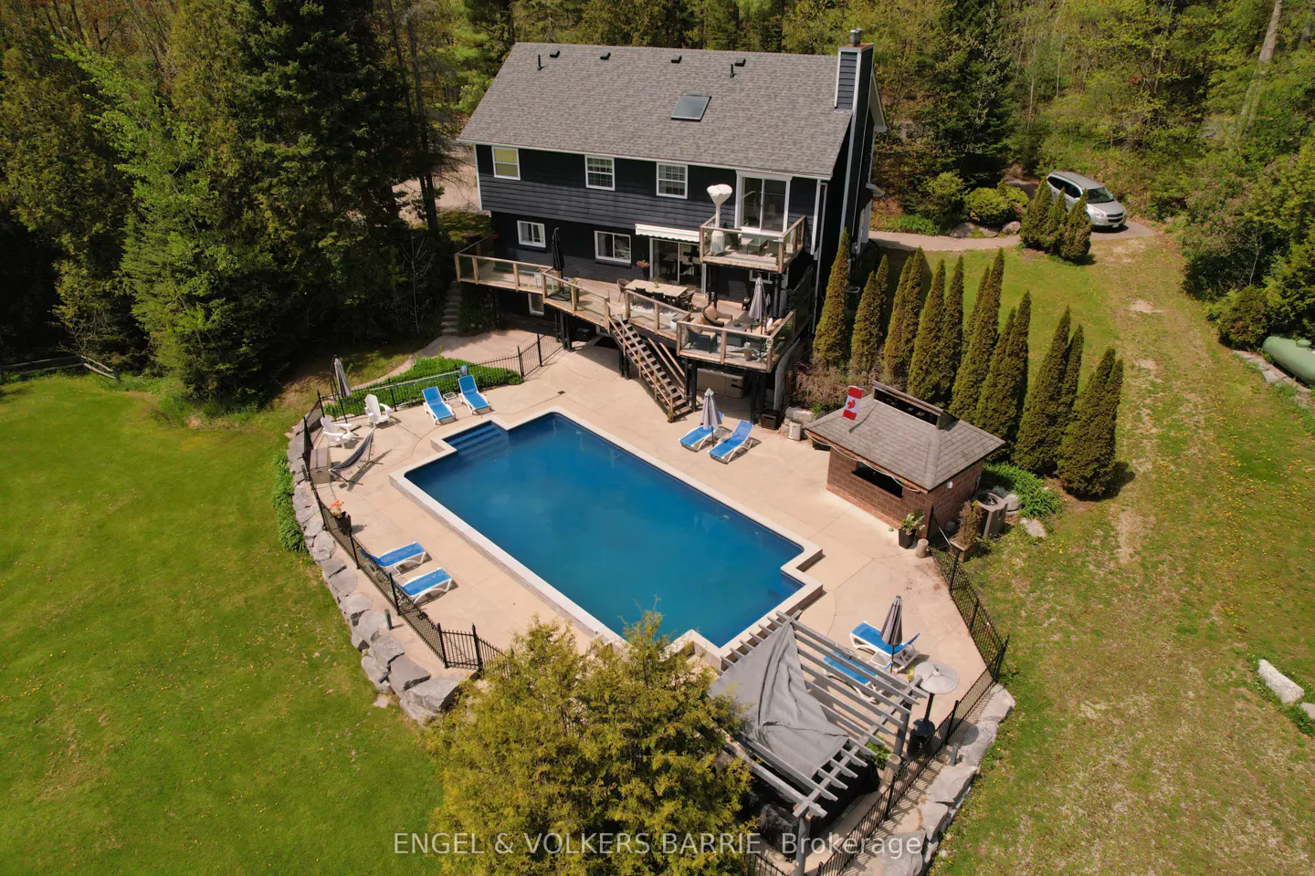 Aerial view of a two-story dark gray house with a pool, deck, and green lawn surrounded by trees.