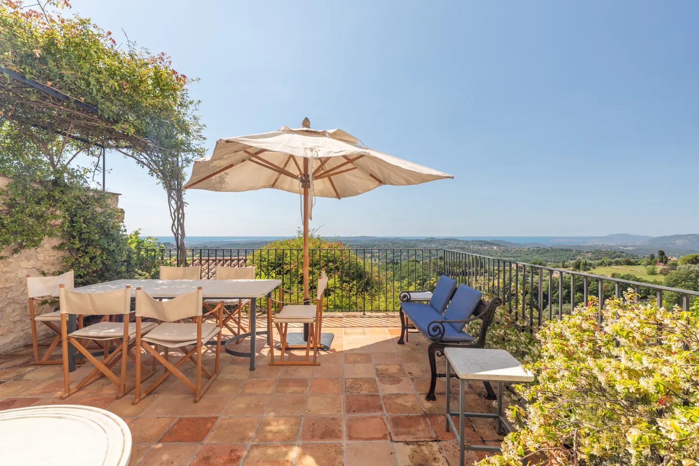 Outdoor patio with table, chairs, umbrella, and blue bench overlooking a green landscape and ocean view.